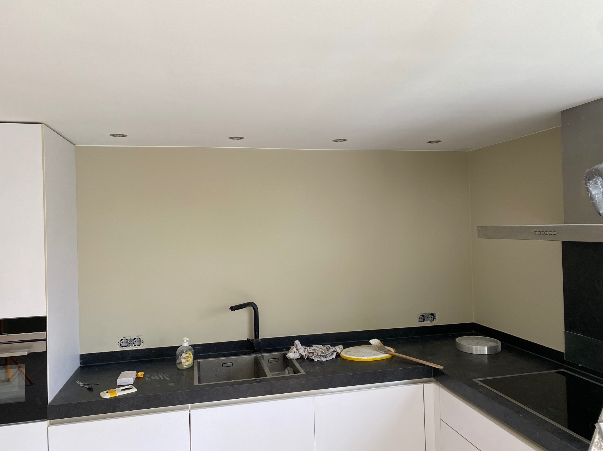 Kuchnia po malowaniu ścian na beżowo, widoczne gniazdka elektryczne, czarny blat, białe szafki i akcesoria malarskie. Oświetlenie sufitowe.