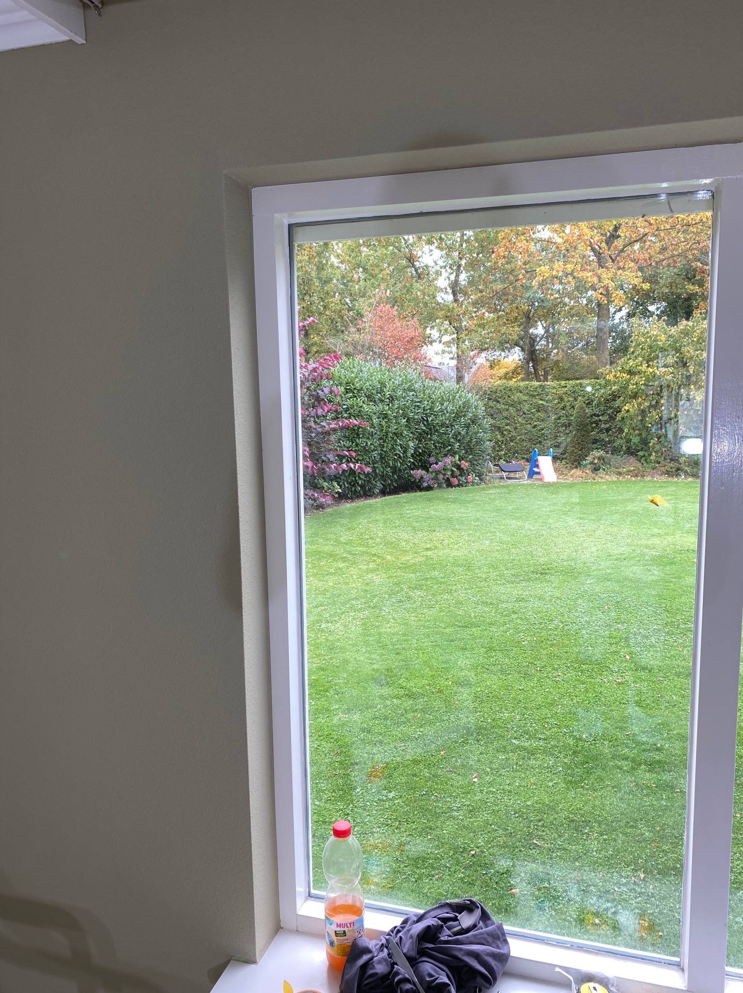 Białe okno z widokiem na zielony ogród z drzewami w jesiennych barwach. Na parapecie butelka soku i ubrania. Ściana w neutralnym odcieniu beżu.