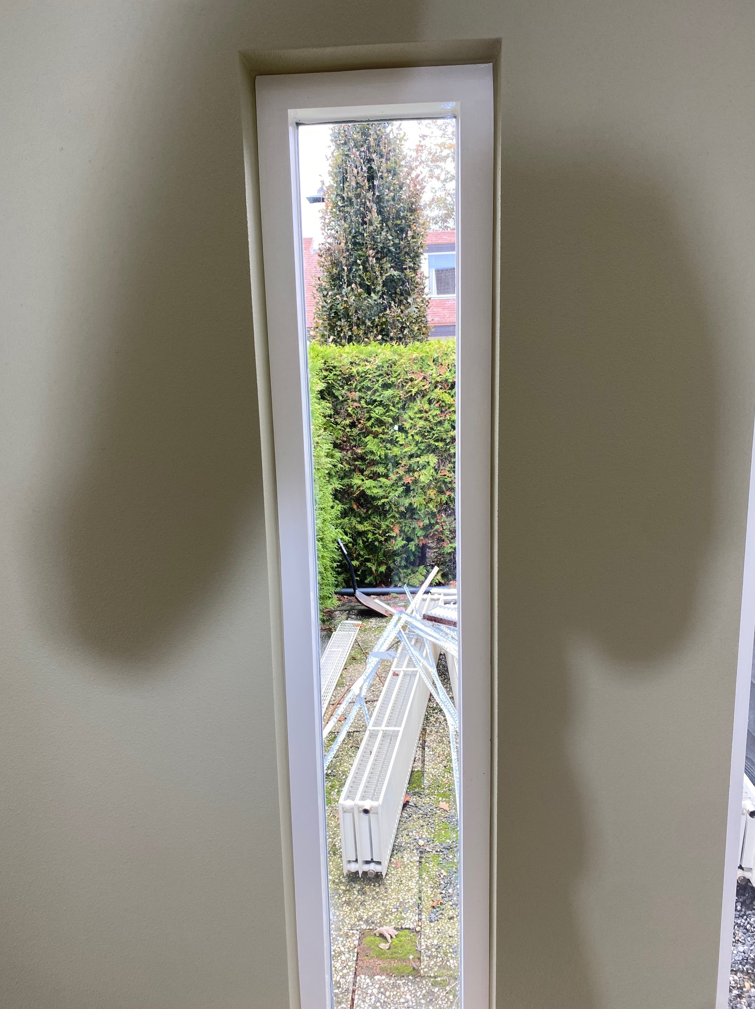 Wąskie okno z białymi ramami w beżowej ścianie, przez które widać ogród z zielenią i fragmentem rusztowania. Widok z wnętrza na zewnątrz.