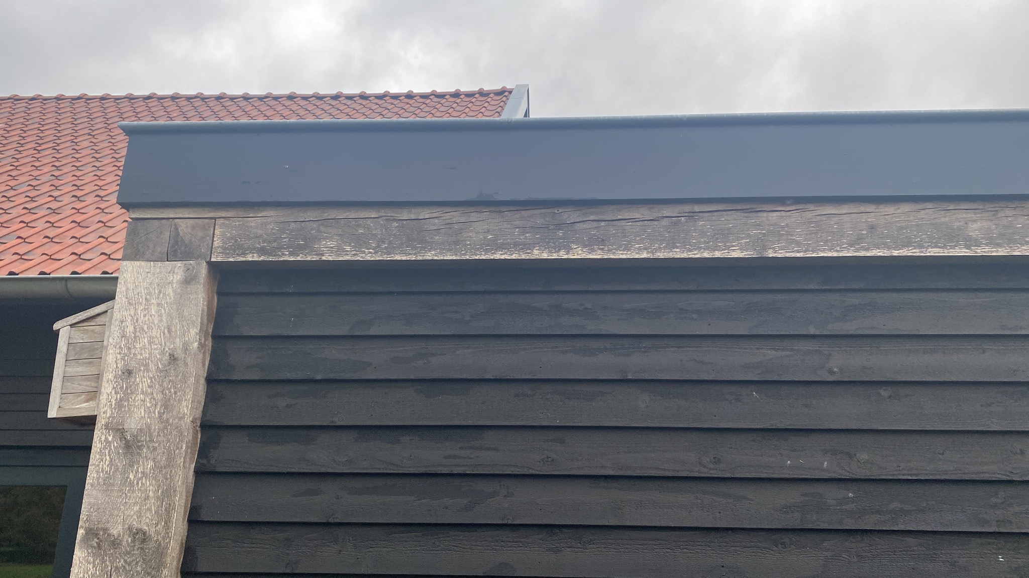 Ciemna, drewniana elewacja budynku z widoczną konstrukcją nośną i fragmentem dachu z czerwonej dachówki w tle. Widoczna faktura drewna i detale wykończenia.