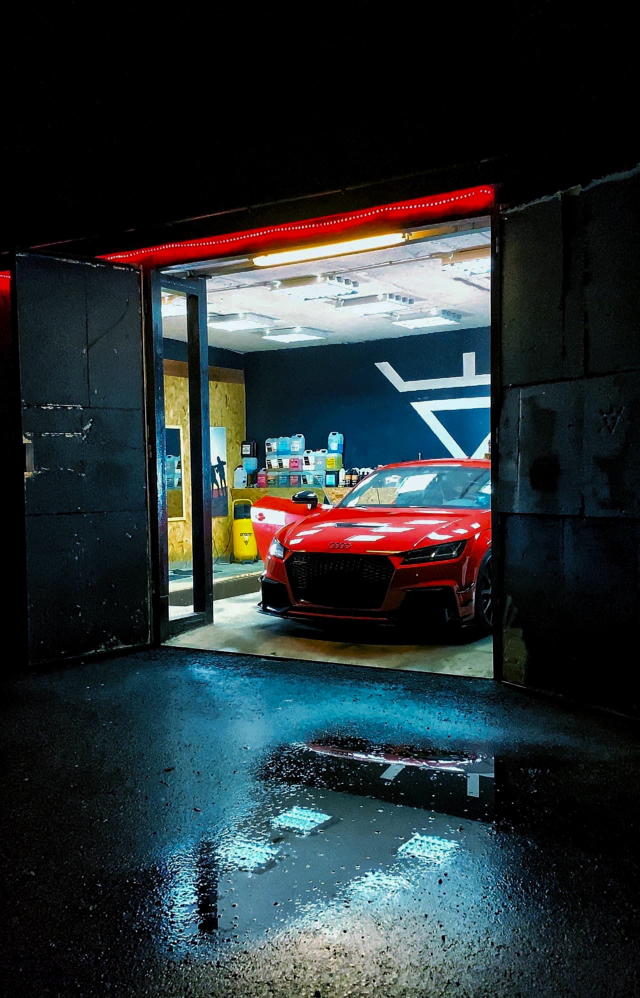 Czerwone Audi TT w myjni z otwartymi drzwiami, mokra posadzka odbija światła. Widoczne kosmetyki samochodowe i logo na ścianie. Ujęcie z zewnątrz, nocne oświetlenie.