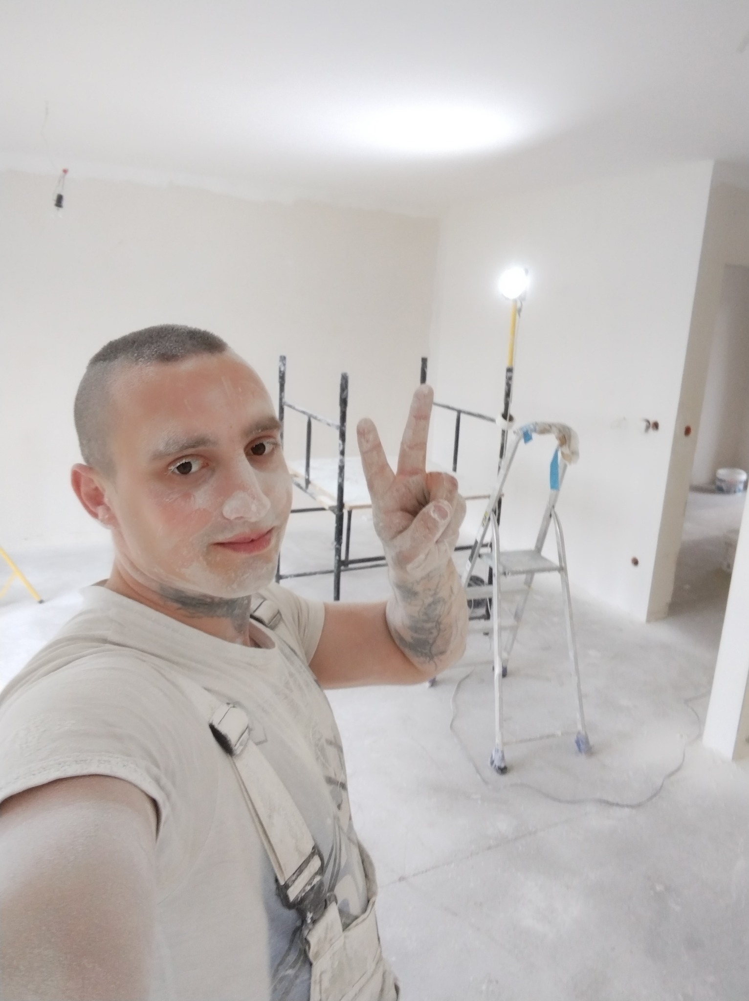 Uśmiechnięty malarz, pokryty pyłem, pokazuje znak zwycięstwa na tle odświeżonego, jasnego wnętrza z rusztowaniem i drabiną. Widoczne ślady po pracy.