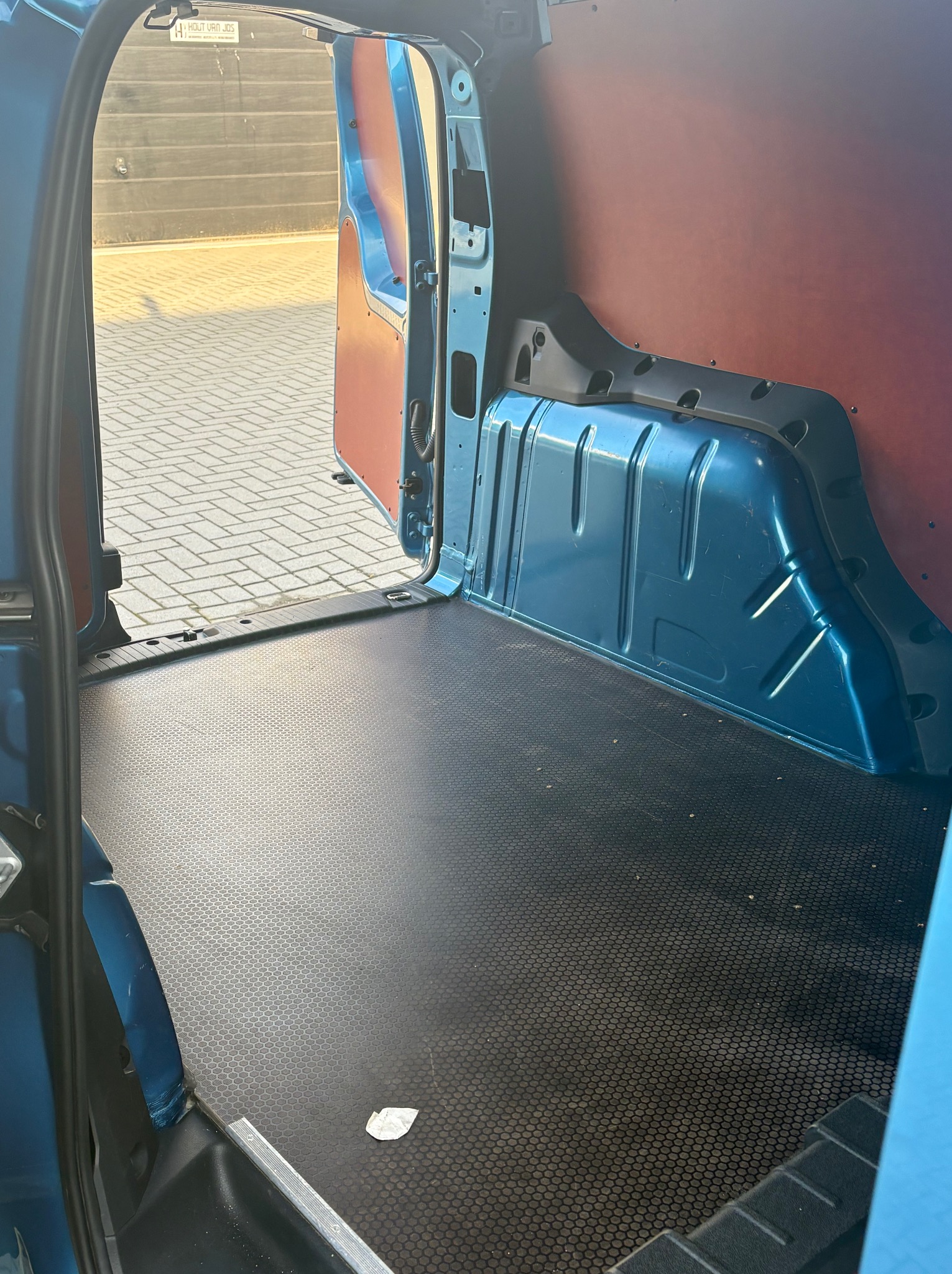 Wnętrze niebieskiego busa z otwartymi drzwiami, widok na pustą przestrzeń ładunkową z czarną podłogą i brązowymi panelami bocznymi. Widoczny fragment chodnika na zewnątrz.