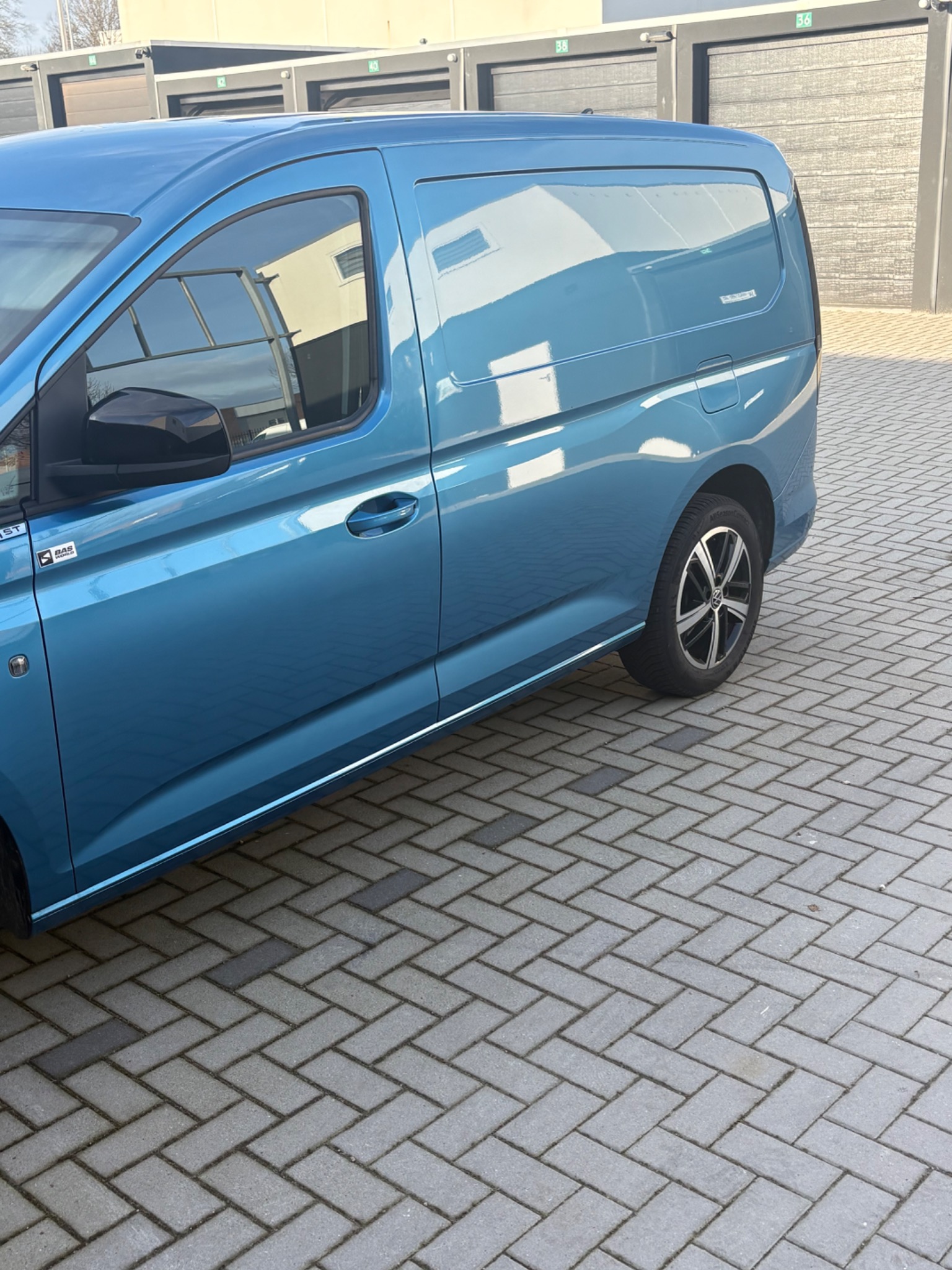 Błękitny bus dostawczy na brukowanym placu, zaparkowany bokiem do kamery, z widocznymi garażami w tle. Czysty, nowoczesny design pojazdu.