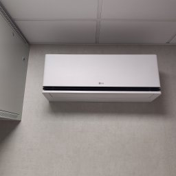 BARTCOMP - Biały klimatyzator LG zamontowany na jasnoszarej, teksturowanej ścianie, obok szarej szafy elektrycznej. Sufit podwieszany z białych paneli akustycznych.