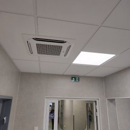 BARTCOMP - Biały klimatyzator kasetonowy LG w podwieszanym suficie, obok panelu LED, w jasnym pomieszczeniu z szarymi ścianami i zielonym znakiem wyjścia ewakuacyjnego nad przeszklonymi drzwiami.