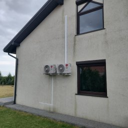 BARTCOMP - Dwie zewnętrzne jednostki klimatyzacji LG Dual Inverter zamontowane na jasnej, teksturowanej ścianie domu jednorodzinnego, z białym orurowaniem biegnącym pionowo. Obok zielony trawnik i chodnik...