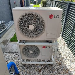 BARTCOMP - Dwie białe jednostki zewnętrzne LG Smart Inverter, zamontowane piętrowo na metalowym stelażu, na jasnym żwirze, obok nowoczesnego ogrodzenia i ciemnej ściany. Widoczna niebieska rura.