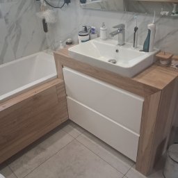 Kam-Bud - Nowoczesna łazienka z wanną obudowaną drewnem i szafką pod umywalką z białymi frontami. Marmurowe płytki na ścianach, szara podłoga. Minimalistyczny design.