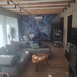 Kam-Bud - Salon z drewnianym sufitem belkowym, ceglaną ścianą i tapetą w marmurkowy wzór. Widoczny szary narożnik, drewniany stolik kawowy i oświetlenie szynowe.