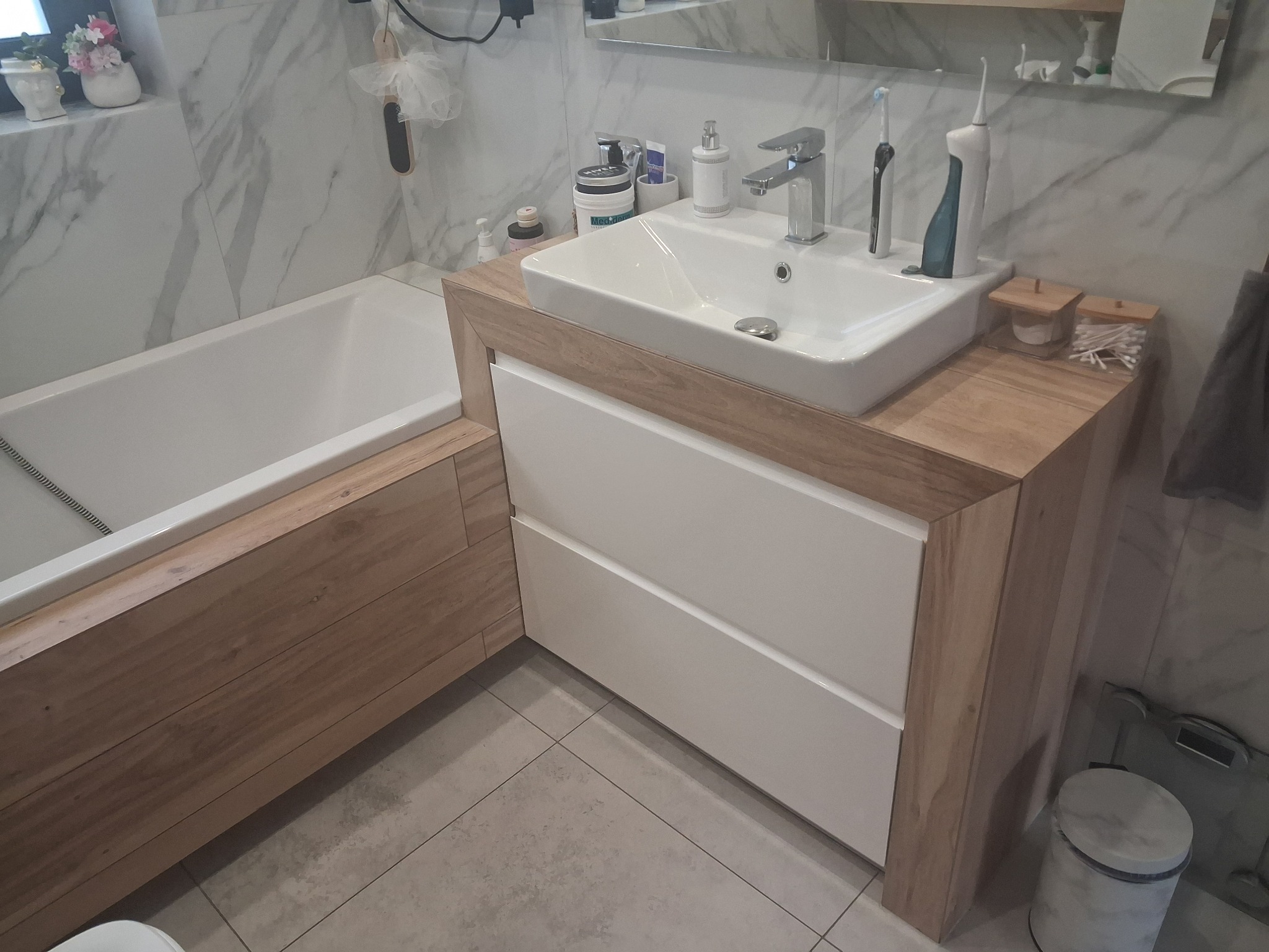 Nowoczesna łazienka z wanną obudowaną drewnem i szafką pod umywalką z białymi frontami. Marmurowe płytki na ścianach, szara podłoga. Minimalistyczny design.