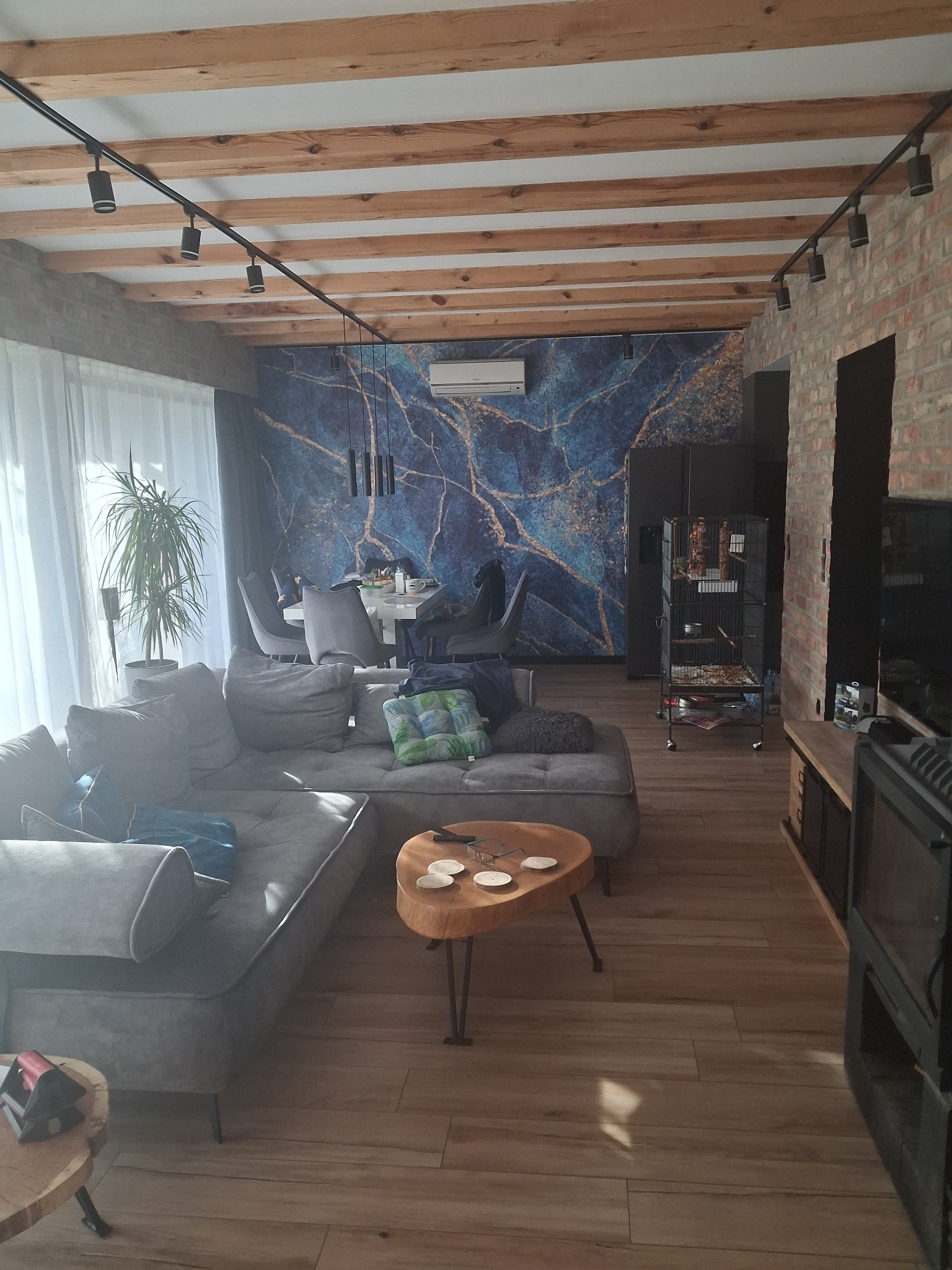 Salon z drewnianym sufitem belkowym, ceglaną ścianą i tapetą w marmurkowy wzór. Widoczny szary narożnik, drewniany stolik kawowy i oświetlenie szynowe.