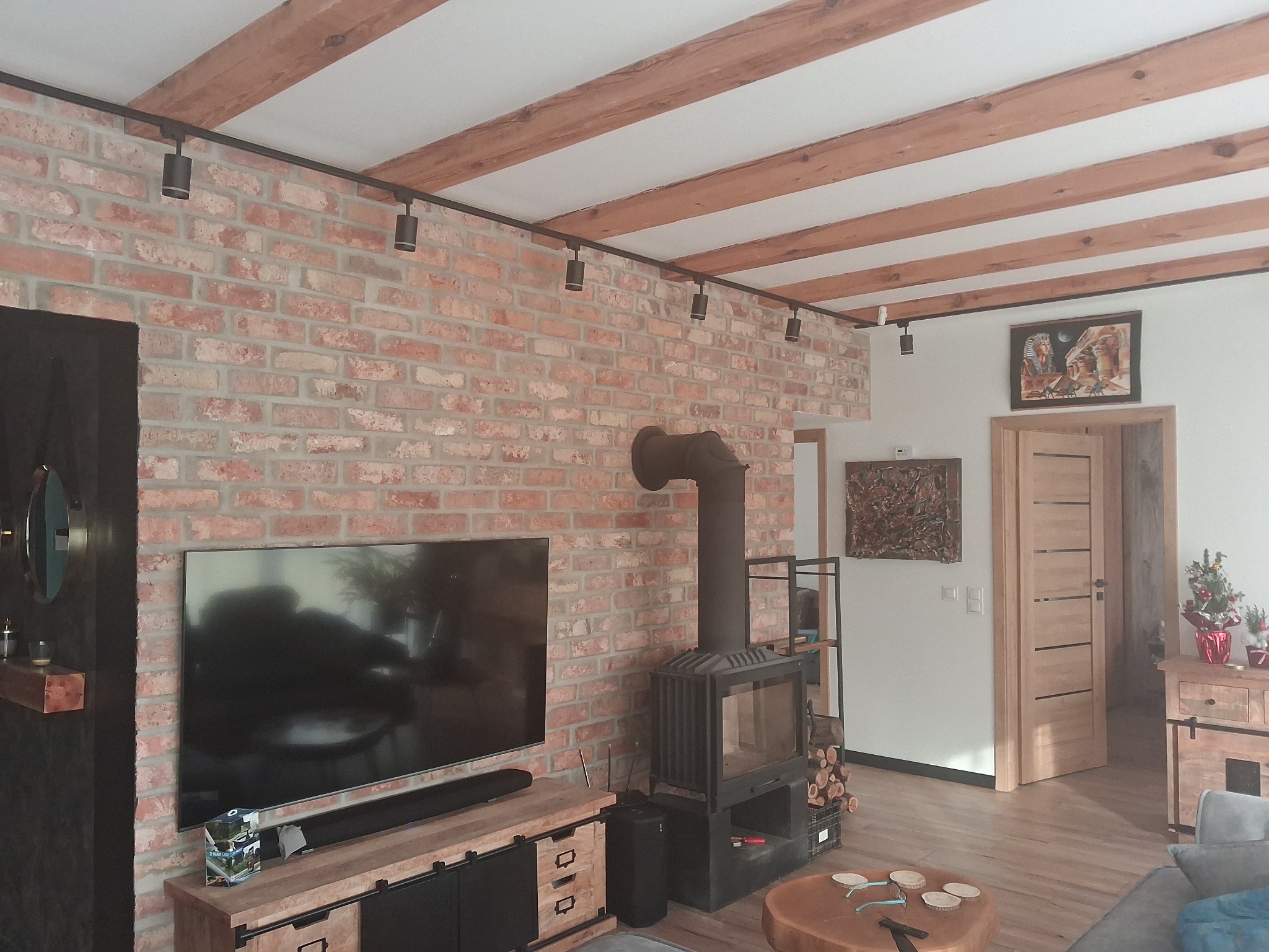 Salon z ceglaną ścianą, drewnianymi belkami stropowymi i kominkiem. Oświetlenie szynowe podkreśla rustykalny charakter wnętrza. Widoczny telewizor i drewniane meble.