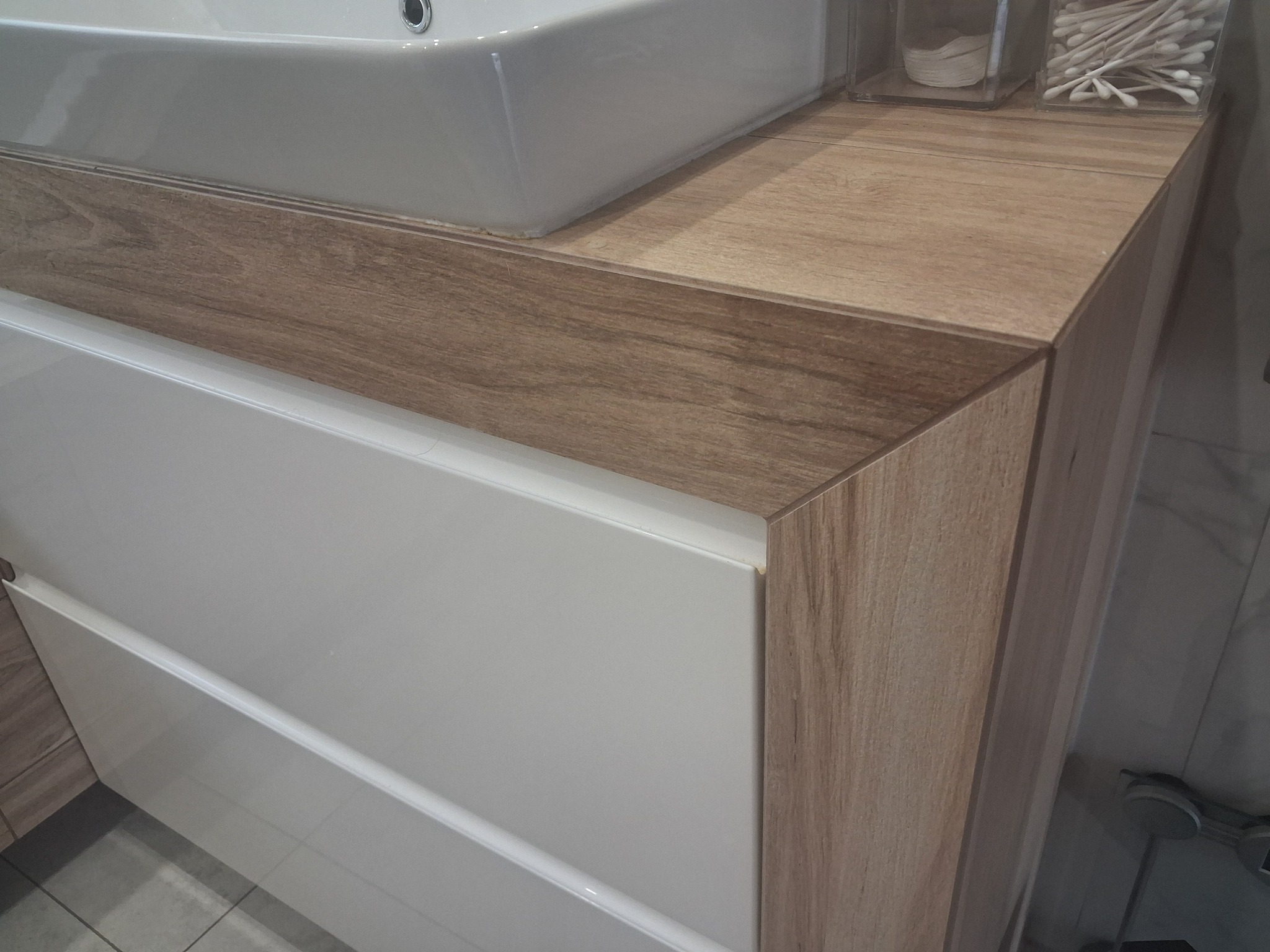 Nowoczesna szafka pod umywalkę z białymi frontami i blatem imitującym drewno. Widoczne detale wykończenia i pojemnik z wacikami.