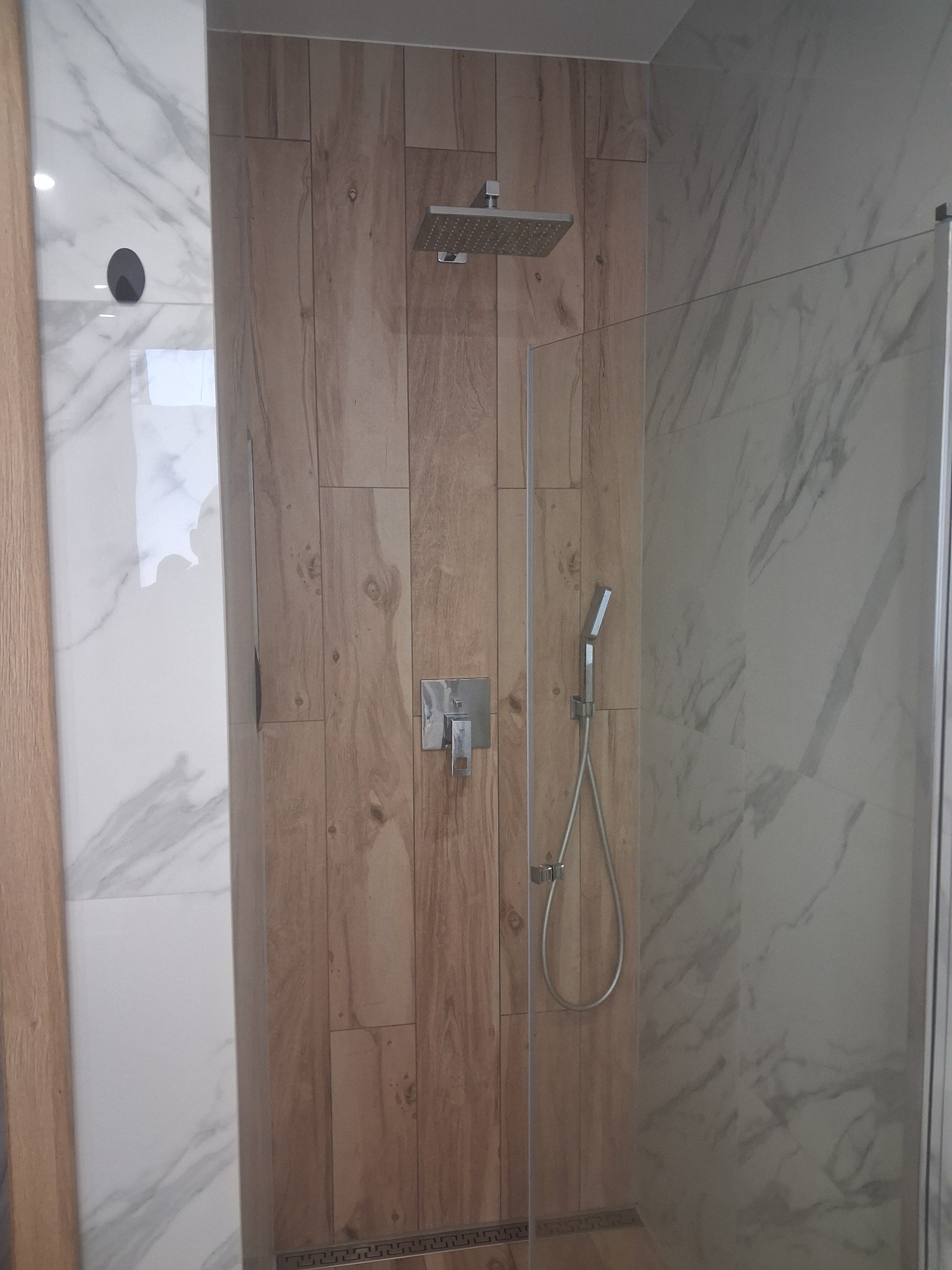 Nowoczesna kabina prysznicowa z drewnopodobnymi i marmurowymi płytkami, deszczownicą i szklaną ścianką. Minimalistyczny design, eleganckie wykończenie.