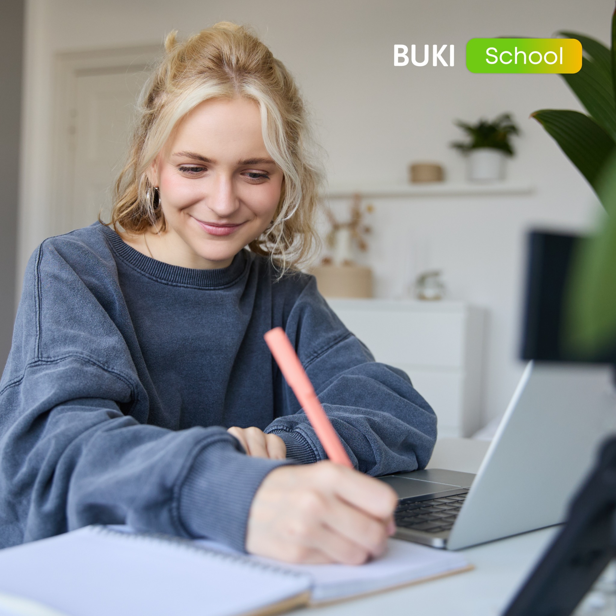 Blondynka pisze w notatniku podczas lekcji online. Widoczny laptop, roślina doniczkowa i logo BUKI School w prawym górnym rogu. Jasne, naturalne światło.