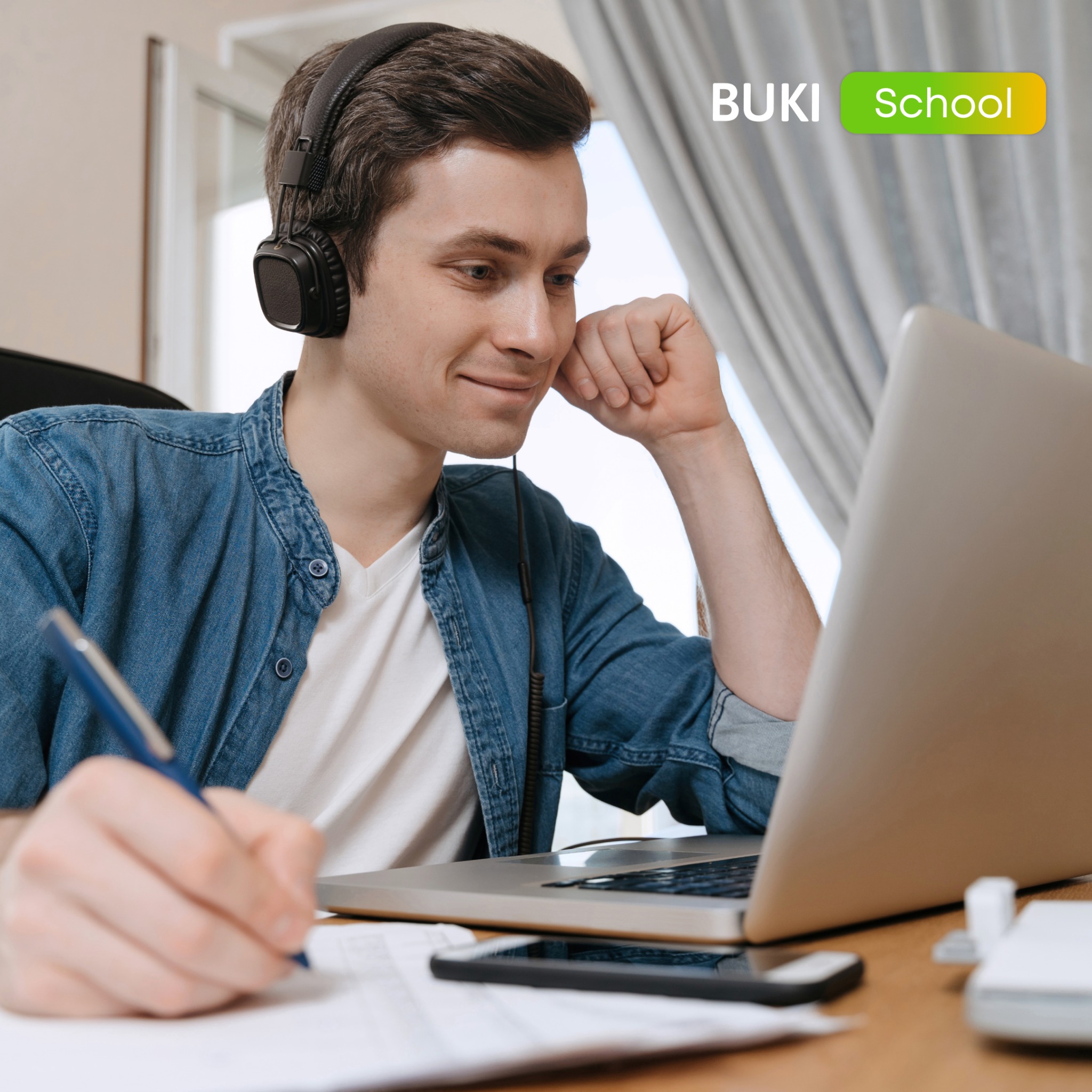 Młody mężczyzna w słuchawkach, uśmiechnięty, pisze notatki podczas lekcji online na laptopie. Logo BUKI School w prawym górnym rogu.