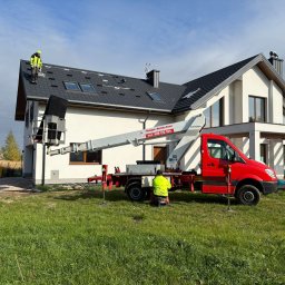 SUN IMPACT SYSTEMY FOTOWOLTAICZNE PIOTR WIŚNIEWSKI - Praca na dachu domu jednorodzinnego z użyciem podnośnika koszowego. Dwóch pracowników w odblaskowych ubraniach. Dach pokryty czarną dachówką z oknami dachowymi.