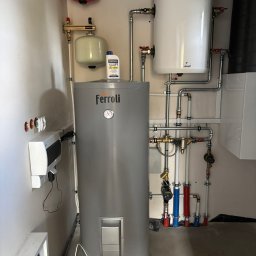 SUN IMPACT SYSTEMY FOTOWOLTAICZNE PIOTR WIŚNIEWSKI - Szary bojler Ferroli z podłączonymi rurami, zbiornik wyrównawczy i biały bojler Galmet na ścianie. Widoczne elementy instalacji grzewczej w kotłowni.