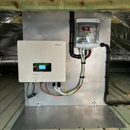 SUN IMPACT SYSTEMY FOTOWOLTAICZNE PIOTR WIŚNIEWSKI - Zamontowany inwerter SOFAR Solar z rozdzielnicą na metalowej płycie w pomieszczeniu z izolacją. Widoczne kable i przewody. Instalacja fotowoltaiczna w trakcie montażu.