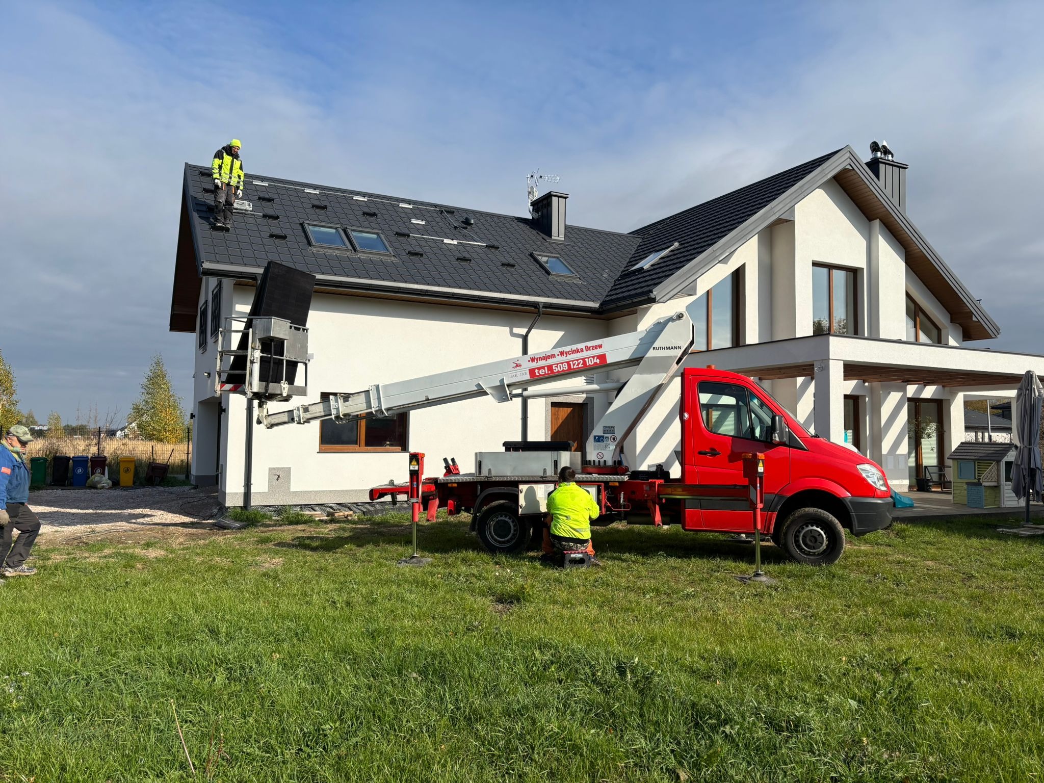 Praca na dachu domu jednorodzinnego z użyciem podnośnika koszowego. Dwóch pracowników w odblaskowych ubraniach. Dach pokryty czarną dachówką z oknami dachowymi.
