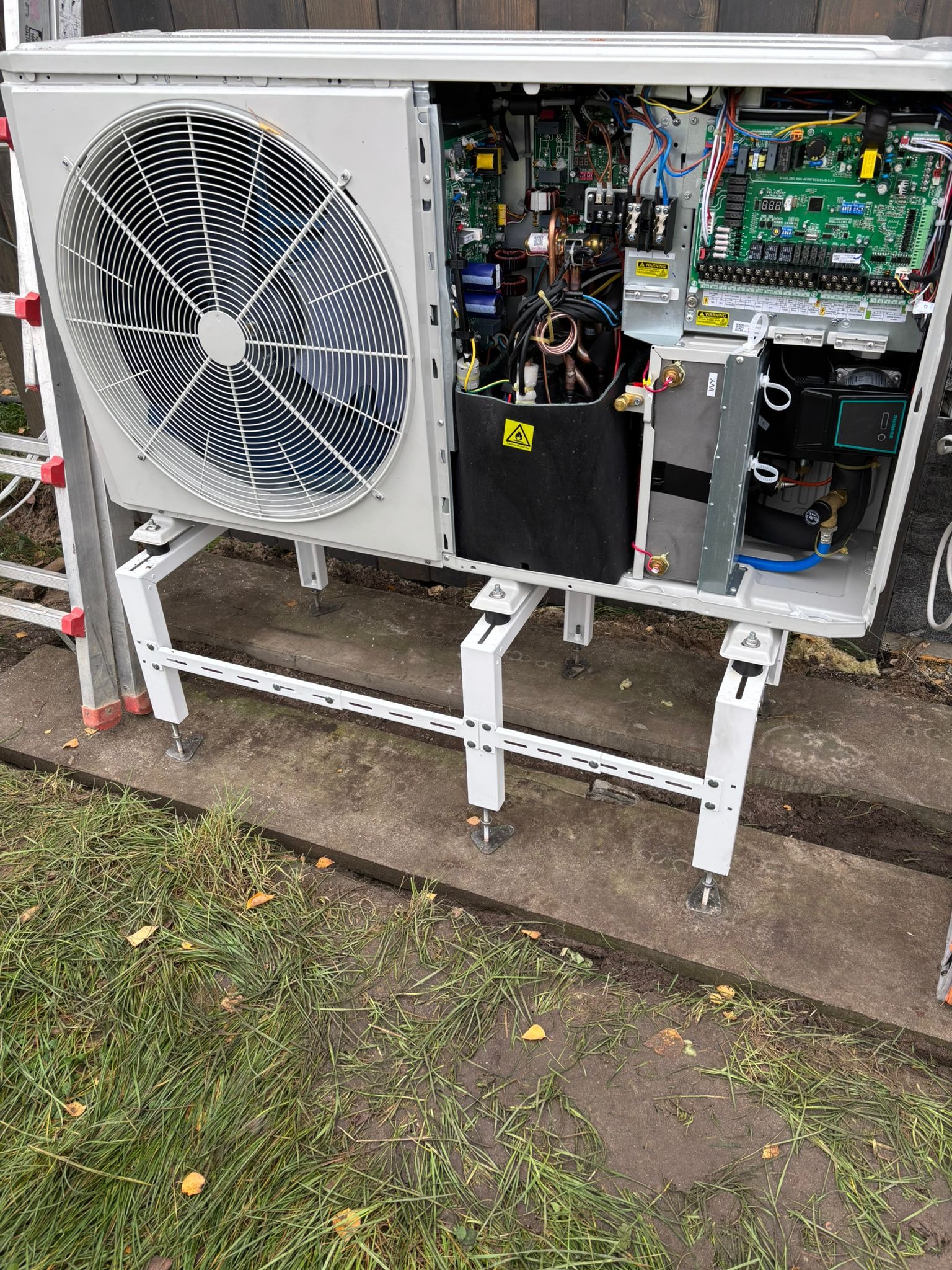 Otwarta obudowa pompy ciepła na białym stelażu, widoczne wewnętrzne komponenty i elektronika. Obok drabina, w tle trawa i betonowa podstawa.
