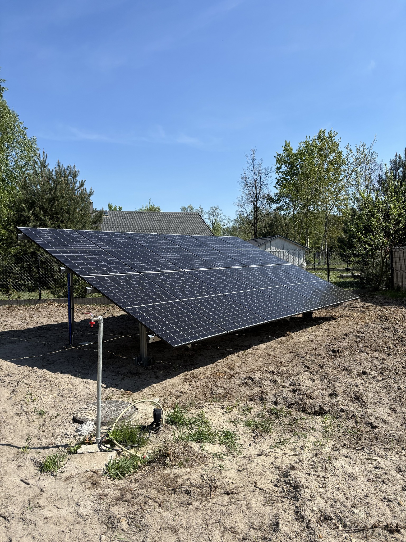 Panel słoneczny na gruncie, widok z boku, instalacja w ogrodzie, błękitne niebo, dom w tle. Widoczne elementy montażowe i przyłącza.