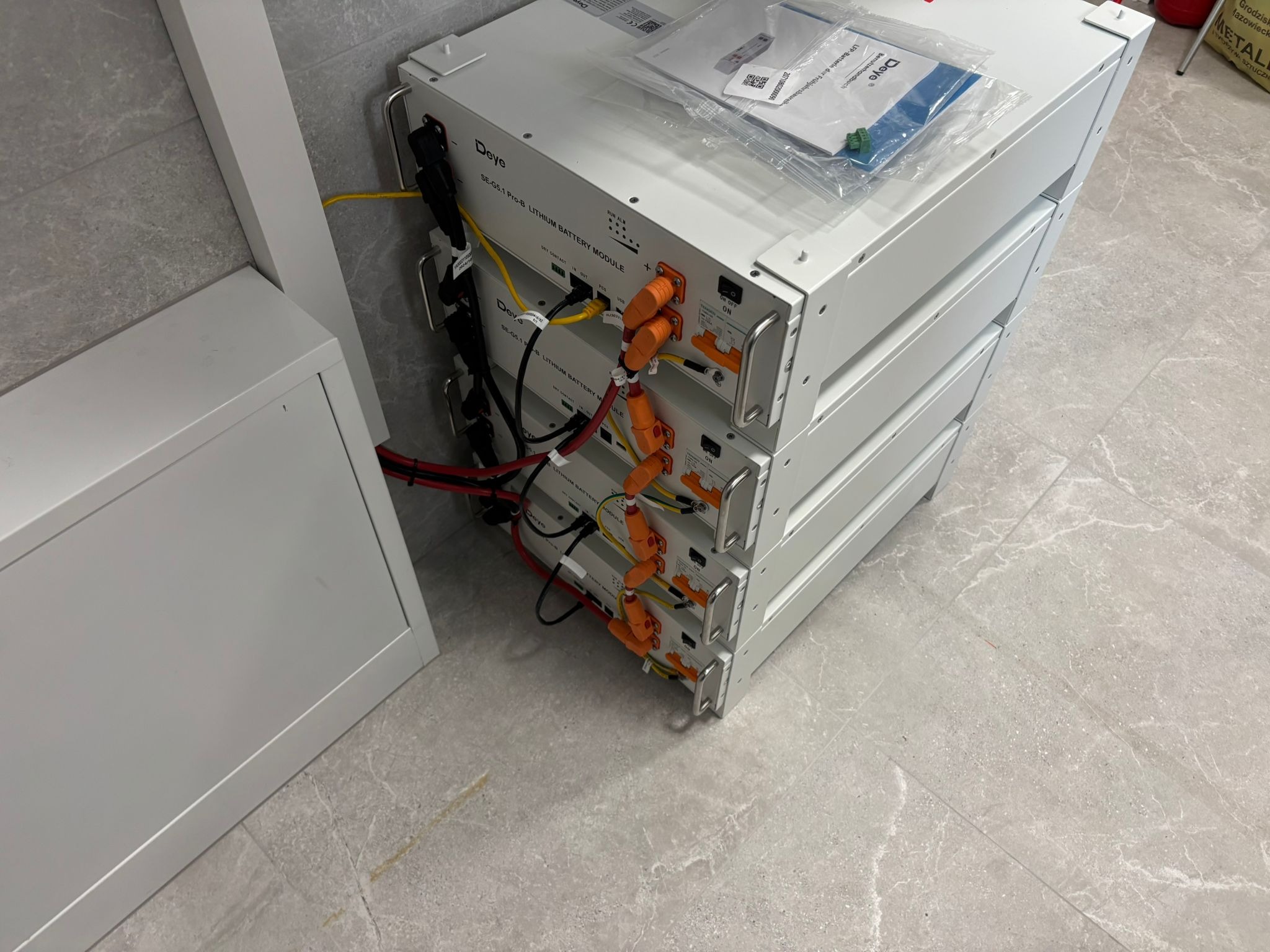 Białe moduły bateryjne Deye SE-G5.1 Pro-B, ułożone jeden na drugim, z widocznymi kablami i złączami, na tle szarej podłogi. Dokumentacja na górnym module.