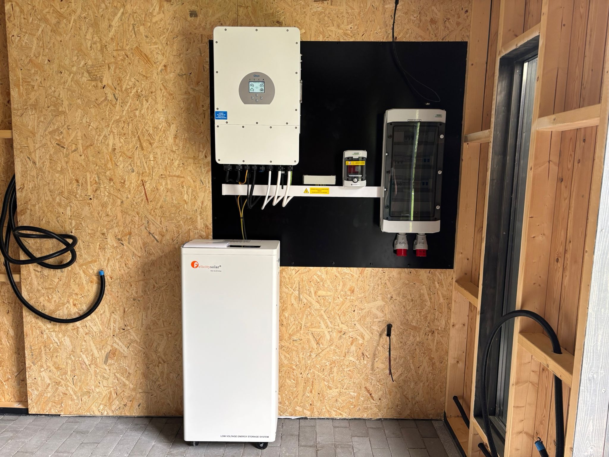 Biały inwerter solarny i magazyn energii Felicity Solar zamontowane na ścianie z płyty OSB w pomieszczeniu w trakcie wykańczania. Widoczne kable i elementy instalacji.