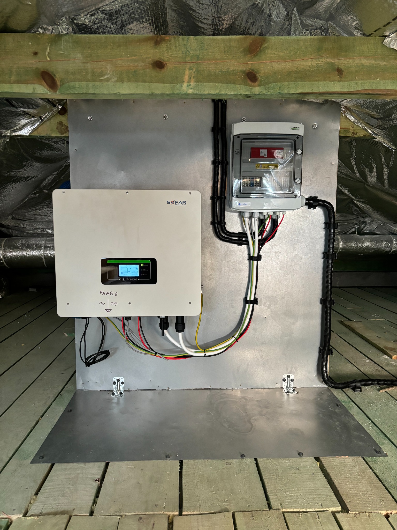 Zamontowany inwerter SOFAR Solar z rozdzielnicą na metalowej płycie w pomieszczeniu z izolacją. Widoczne kable i przewody. Instalacja fotowoltaiczna w trakcie montażu.