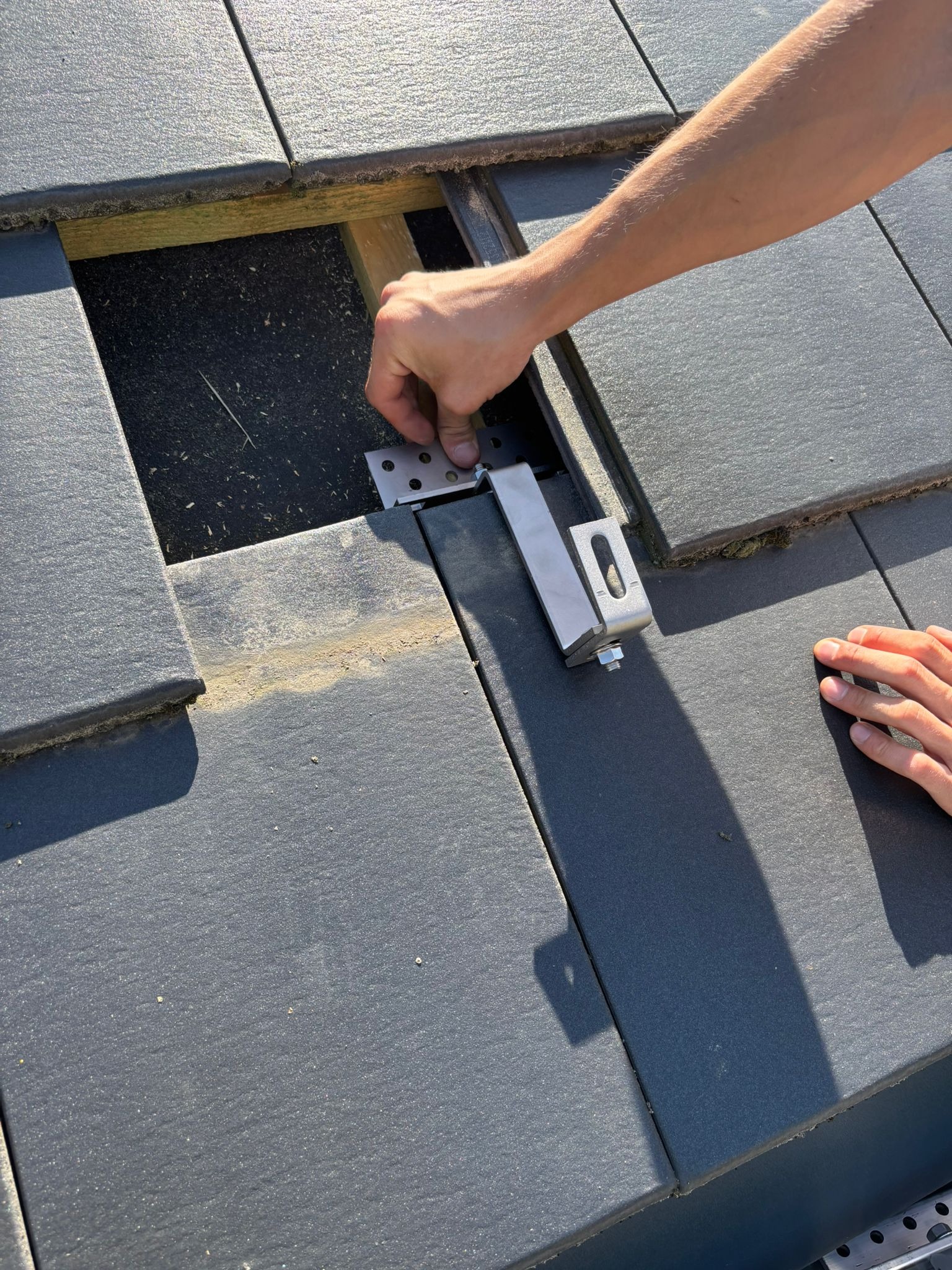 Montaż uchwytu na dachu z szarej dachówki. Widoczne ręce instalatora mocujące element. Otwór w dachu odsłania konstrukcję drewnianą. Jasne oświetlenie.