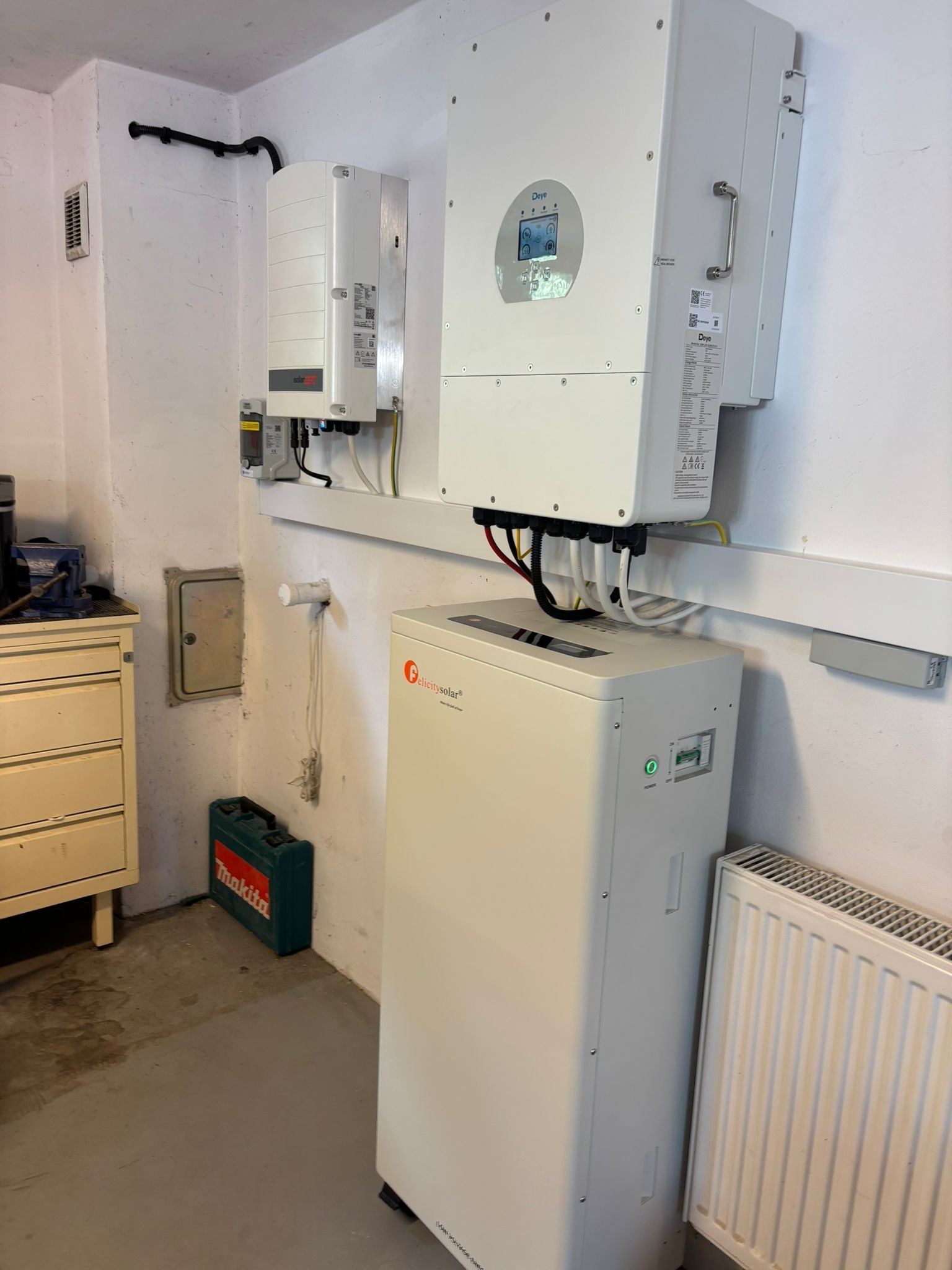 Biały akumulator energii Felicitysolar i dwa falowniki Deye zamontowane na ścianie w pomieszczeniu. Widoczne kable i rury.