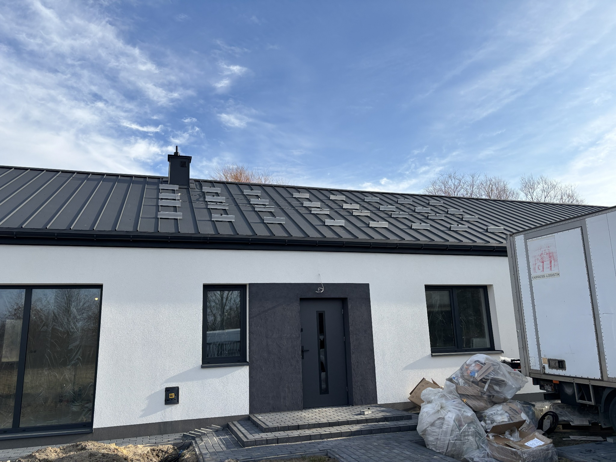 Dom jednorodzinny z szarą blachą na dachu, na którym zamontowano elementy instalacji fotowoltaicznej. Widoczne okna, drzwi wejściowe i fragment ciężarówki z boku budynku.