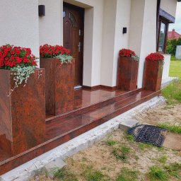 1. ZAKŁAD KAMIENIARSKO - BETONIARSKI RYSZARD JASEK - Kamienne schody wejściowe z donicami z czerwonego granitu, ozdobione czerwonymi kwiatami. Polerowana powierzchnia odbija światło, tworząc elegancki efekt.