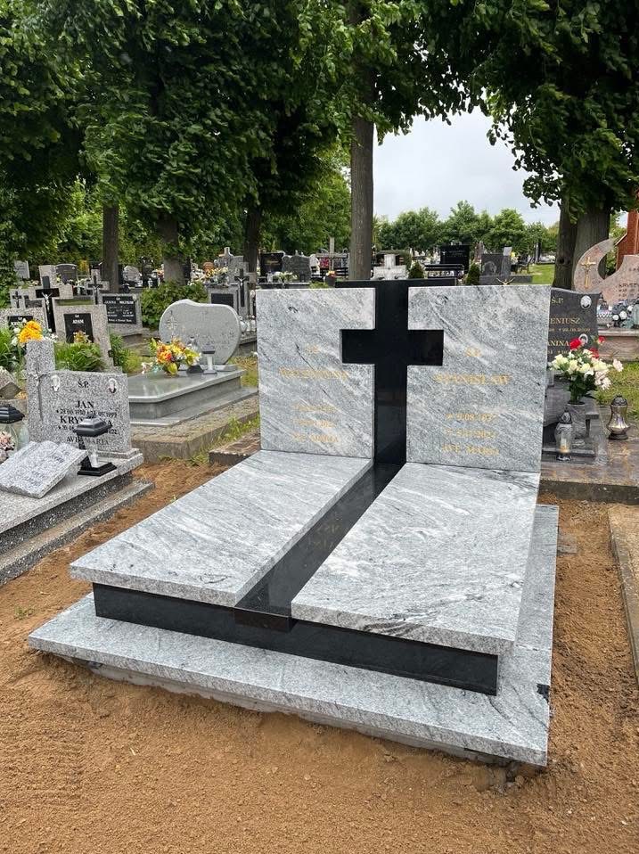 Granitowy nagrobek z czarnym krzyżem na cmentarzu, otoczony zielenią drzew. Jasnoszary kamień z delikatnym wzorem. Widoczne inne groby w tle.