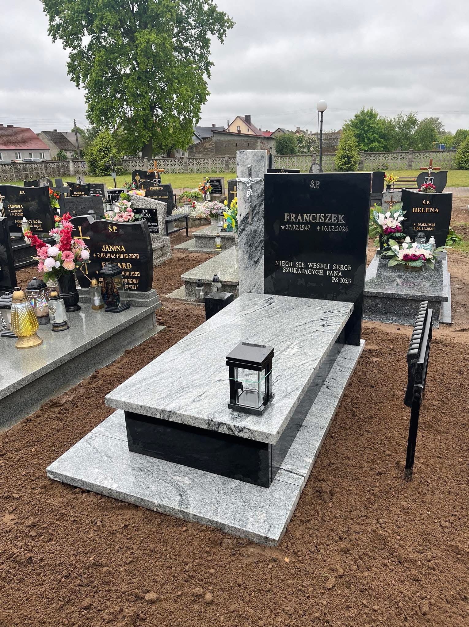 Nowoczesny, dwupoziomowy nagrobek z szarego i czarnego kamienia na cmentarzu, z lampionem. Widoczne inne nagrobki i drzewa w tle. Pochmurne niebo.