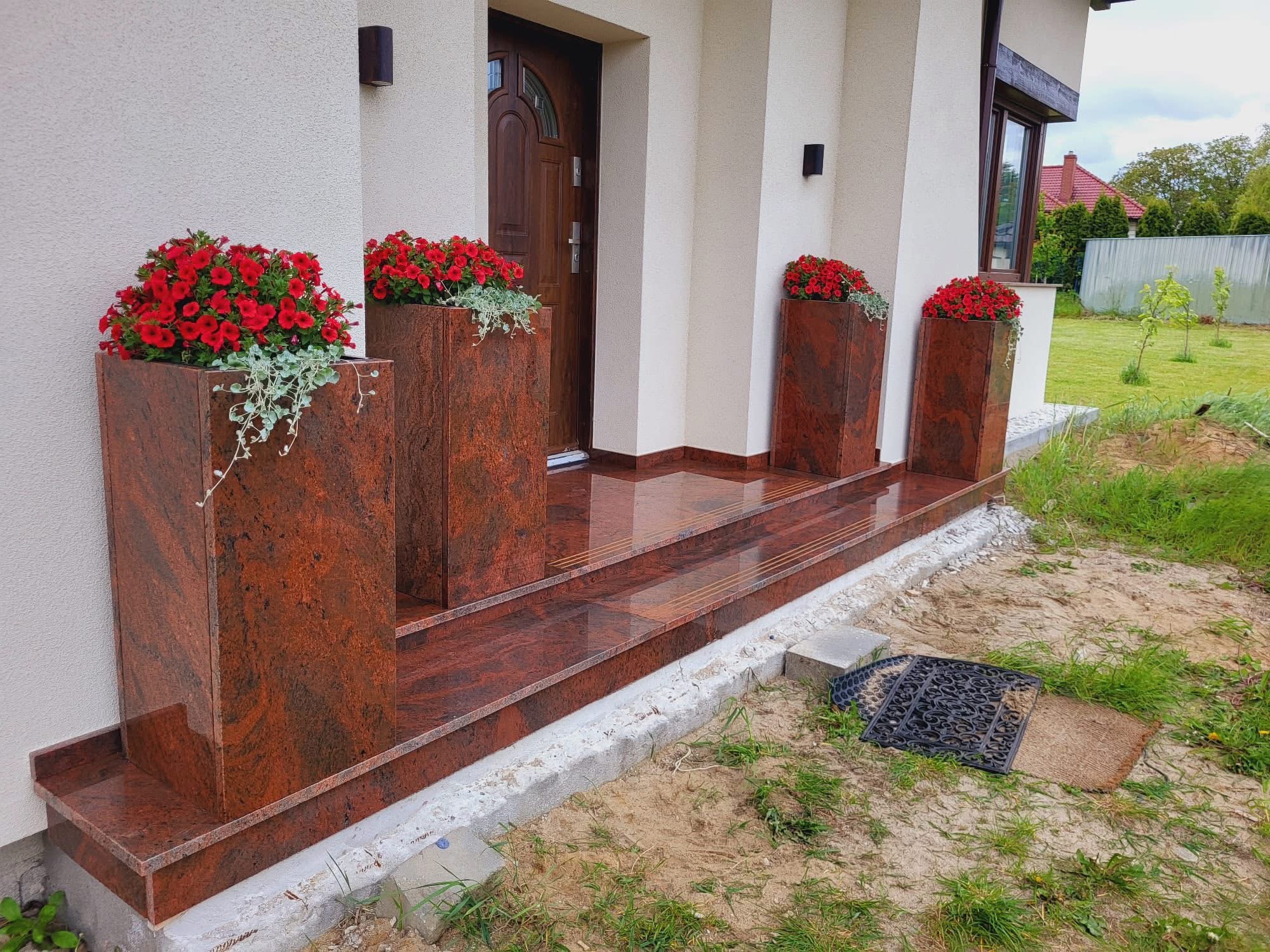 Kamienne schody wejściowe z donicami z czerwonego granitu, ozdobione czerwonymi kwiatami. Polerowana powierzchnia odbija światło, tworząc elegancki efekt.