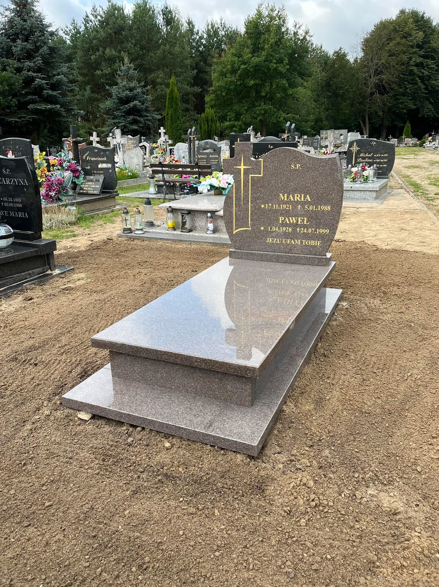 Nowy, brązowo-szary nagrobek z granitu na cmentarzu, z wygrawerowanymi złotymi napisami i krzyżem. Świeżo usypana ziemia wokół pomnika, w tle inne groby i drzewa.