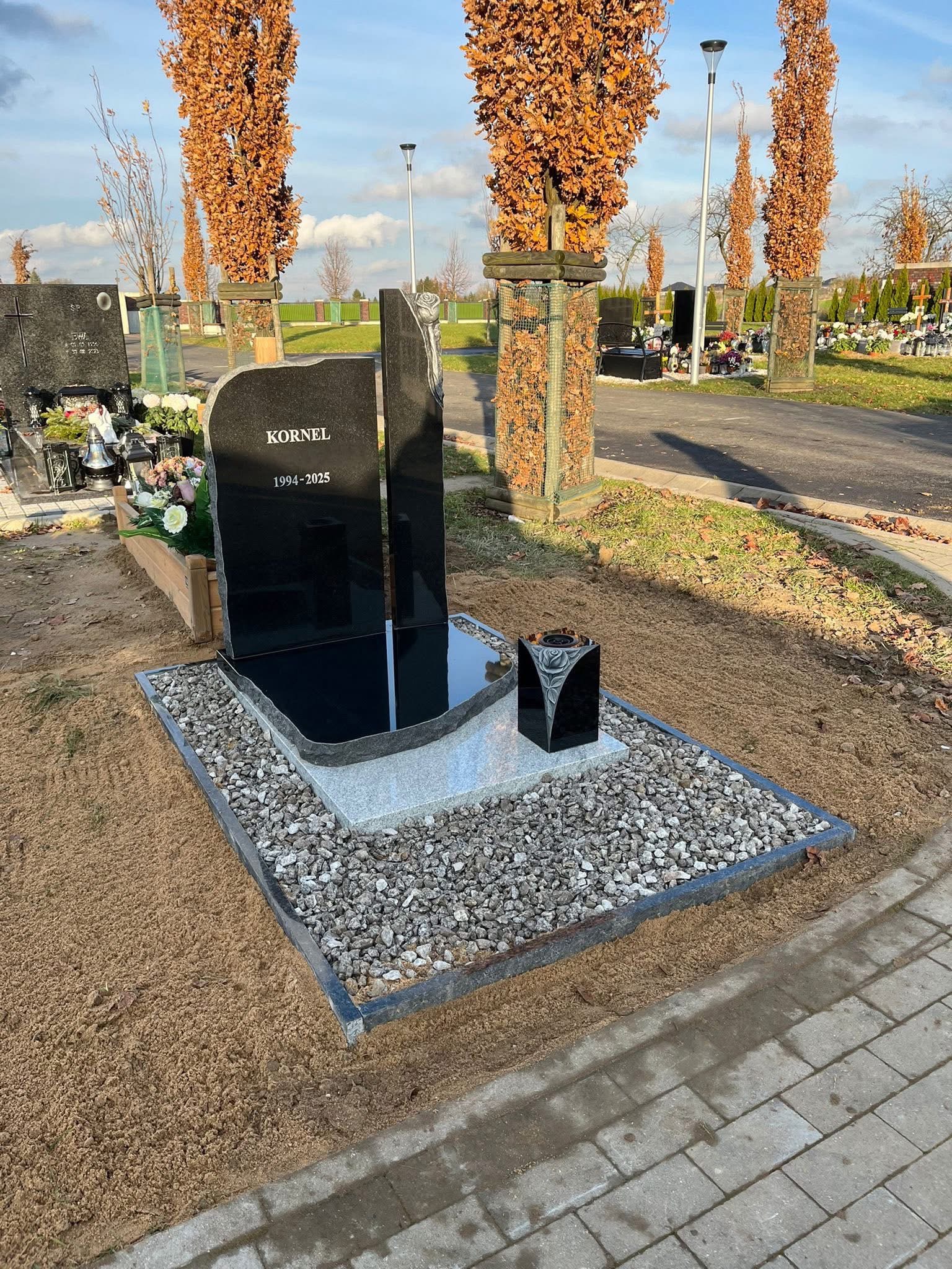 Czarny, nowoczesny nagrobek z wygrawerowanym napisem 'KORNEL 1994-2025' na cmentarzu, otoczony kamieniami i ziemią. W tle drzewa z brązowymi liśćmi.