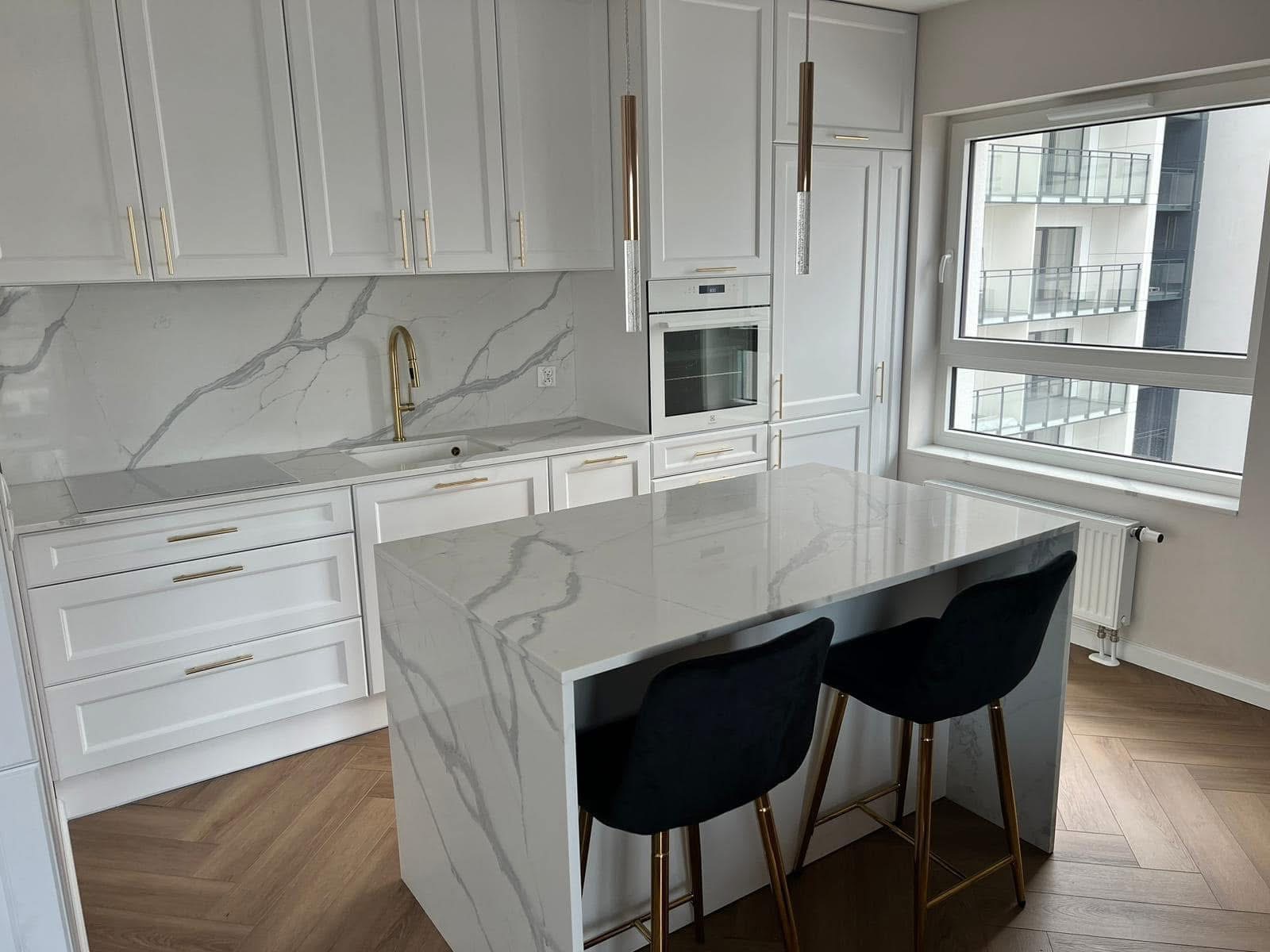 Biała kuchnia z wyspą i blatem z konglomeratu, złote akcenty, nowoczesny design, parkiet w jodełkę. Widok na budynek zza okna.