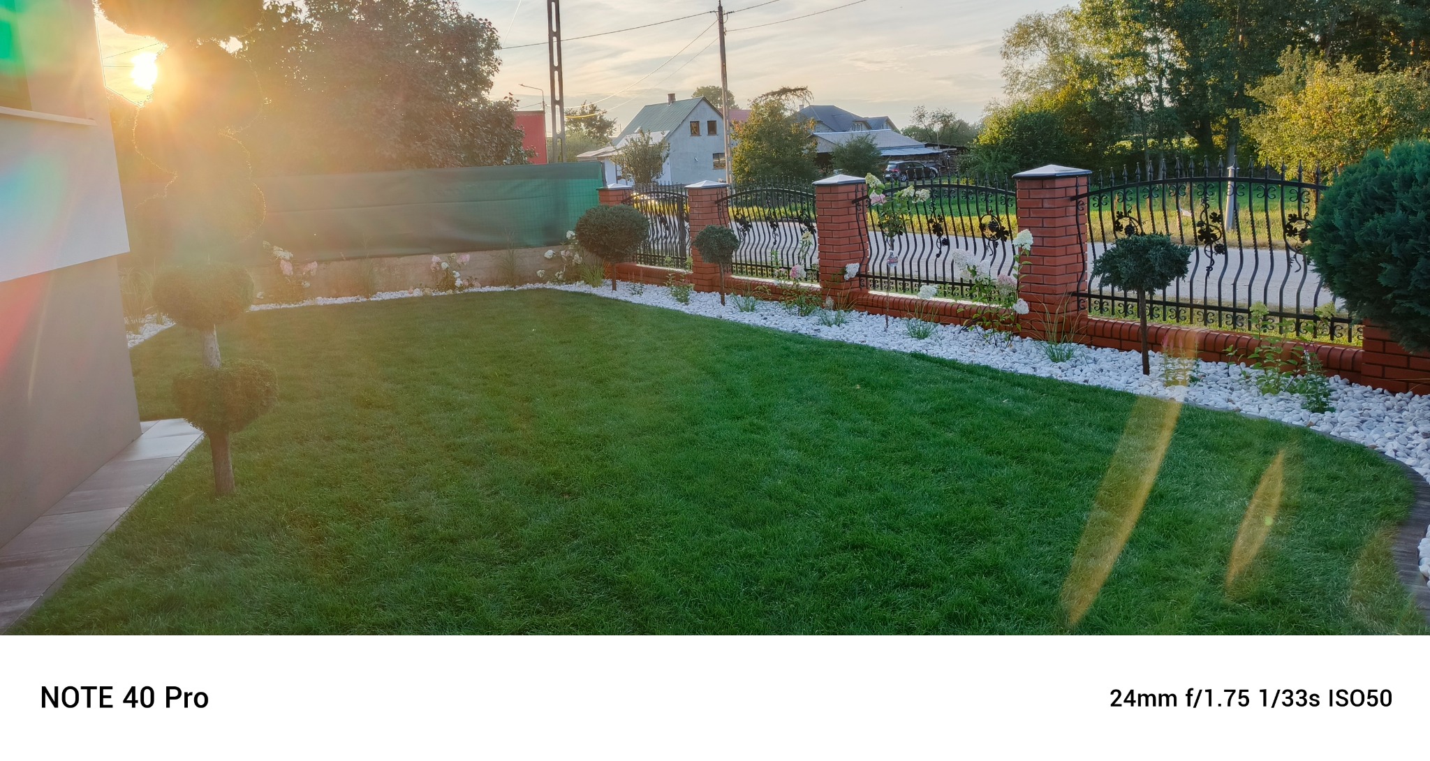 Zadbany trawnik z równo przystrzyżoną trawą, obrzeża z białych kamieni, dekoracyjne krzewy i ozdobne ogrodzenie z ceglanymi słupkami w słoneczny dzień.