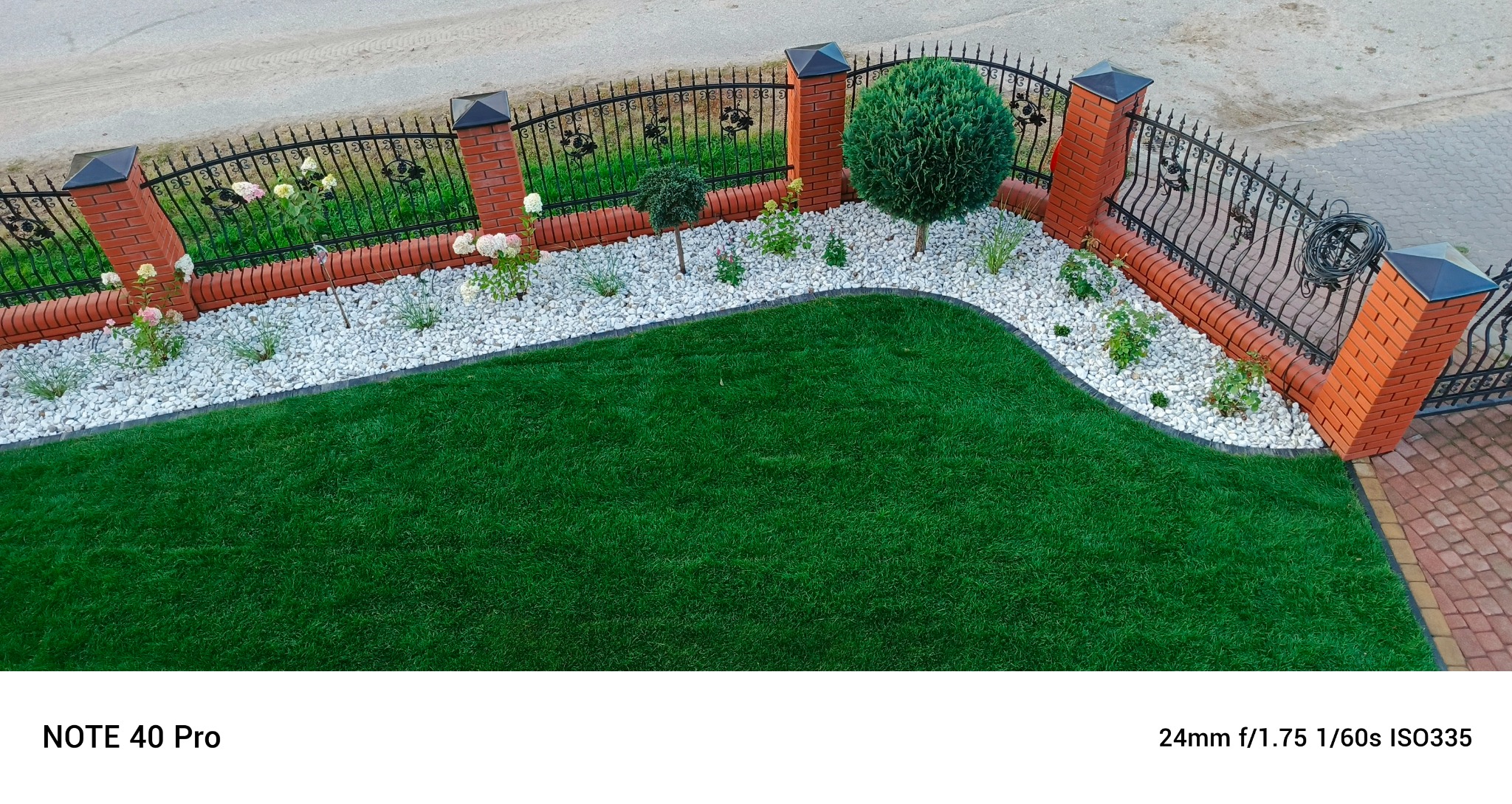 Widok z góry na zadbany ogród z równo przystrzyżonym trawnikiem, białymi kamieniami, niskimi krzewami i ozdobnym ogrodzeniem z ceglanymi słupkami.