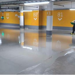 DS-eco Sp zoo - Aplikacja szarej posadzki żywicznej w garażu podziemnym. Dwóch pracowników rozprowadza masę, widoczne numery miejsc parkingowych na żółtych ścianach.