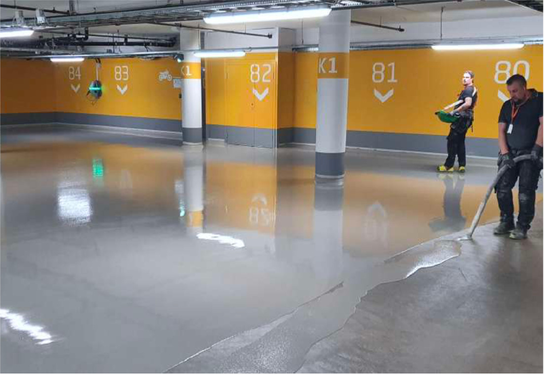 Aplikacja szarej posadzki żywicznej w garażu podziemnym. Dwóch pracowników rozprowadza masę, widoczne numery miejsc parkingowych na żółtych ścianach.