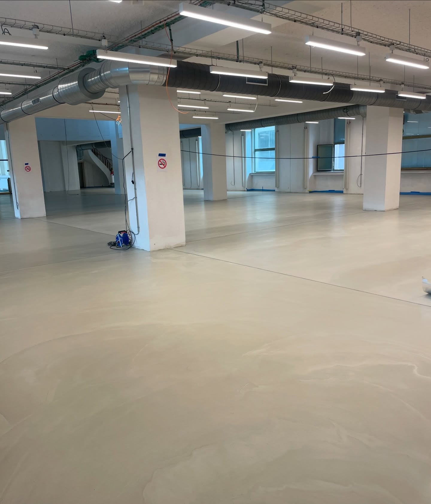 Jasna, gładka posadzka w przestronnym, industrialnym wnętrzu z kolumnami i widocznymi instalacjami pod sufitem. Minimalistyczny design, duże okna w tle.