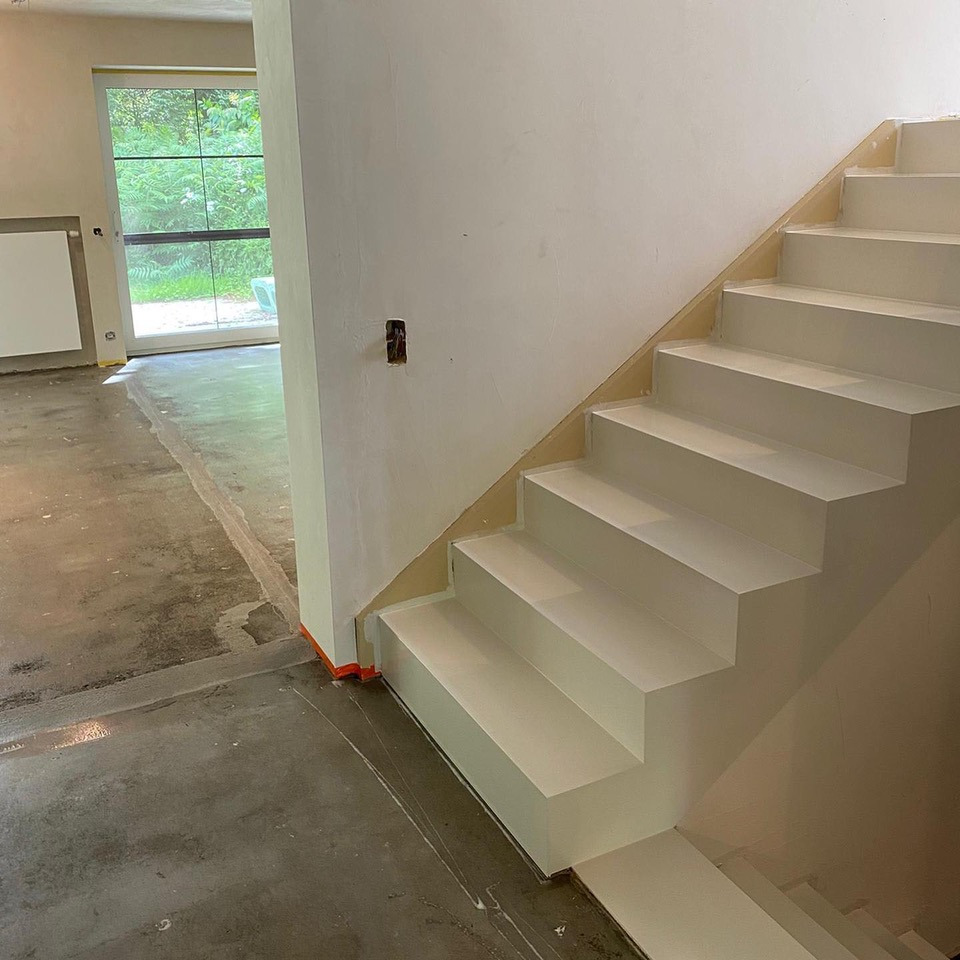 Białe schody w surowym wnętrzu z widoczną wylewką podłogową i oknem. Pomarańczowa taśma zabezpieczająca przy ścianie. Widok na zieleń za oknem.