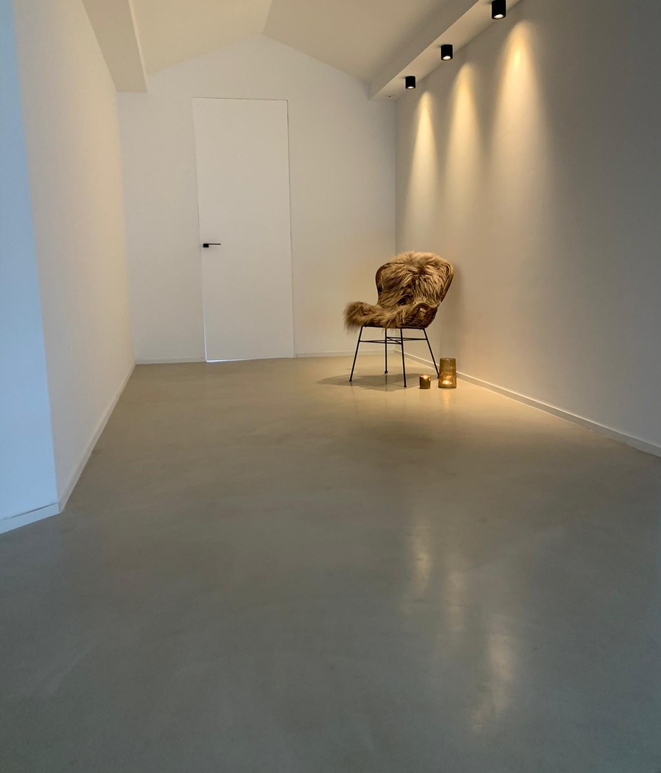 Minimalistyczne wnętrze z gładką, szarą posadzką, fotel z futrzanym okryciem i świecami. Białe ściany i sufit z reflektorami tworzą nowoczesny, jasny klimat.