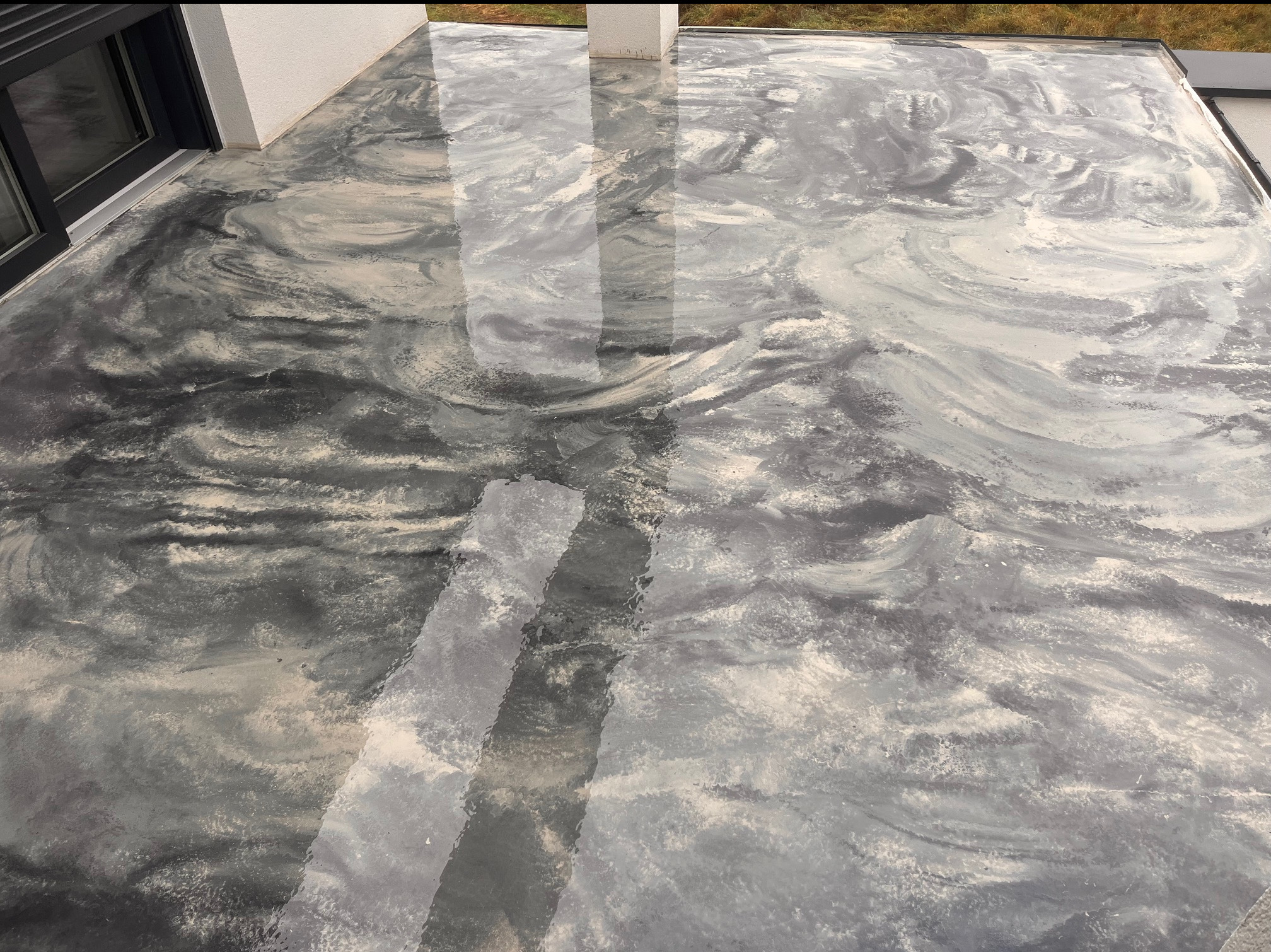 Szara, błyszcząca posadzka żywiczna na tarasie zewnętrznym, z unikalnym wzorem przypominającym marmur, widoczna z góry, z fragmentem okna i słupa w tle.