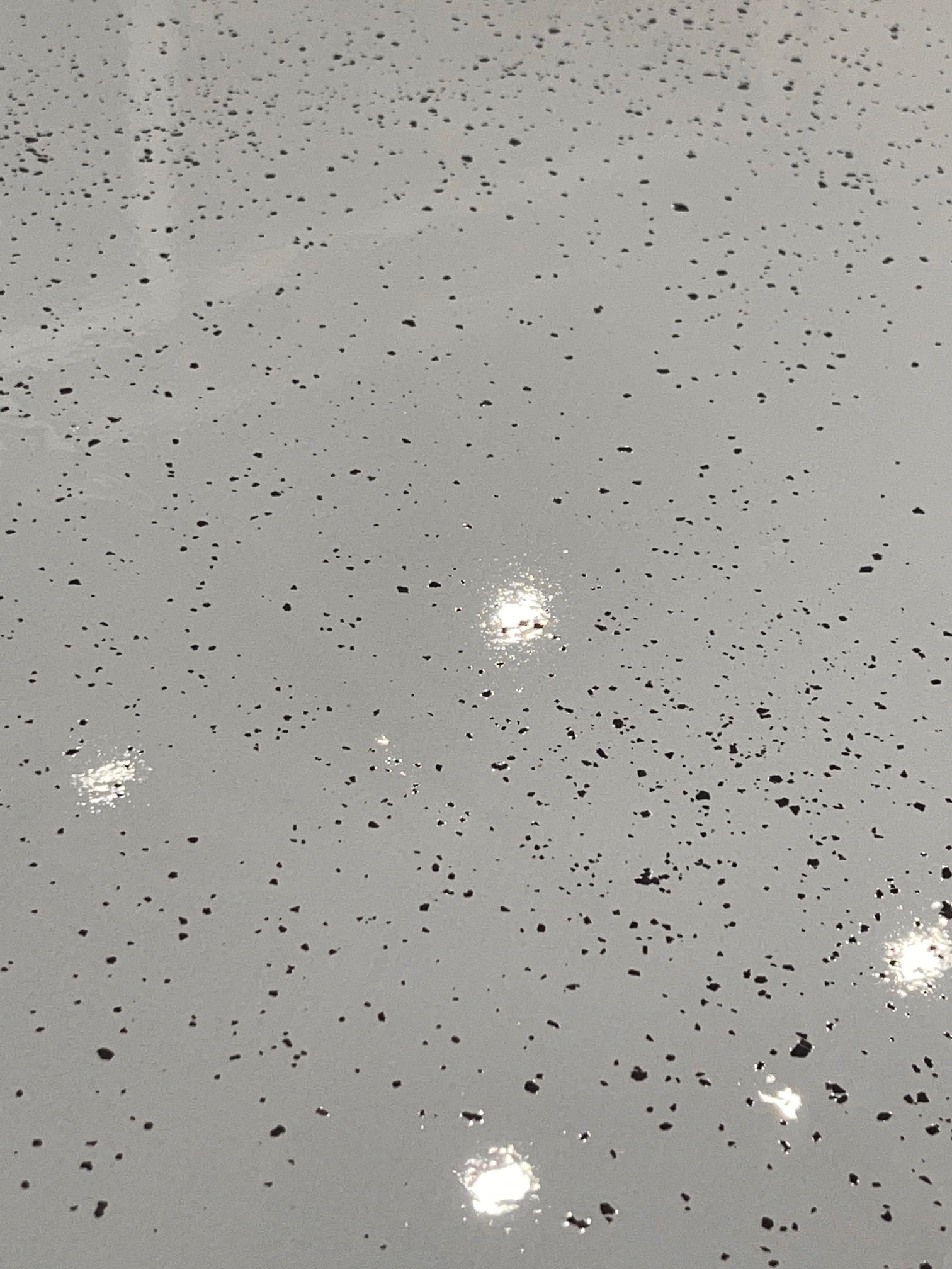 Jasnoszara, gładka powierzchnia z ciemnymi, nieregularnymi plamkami. Widoczne refleksy świetlne podkreślają połysk wykończenia.