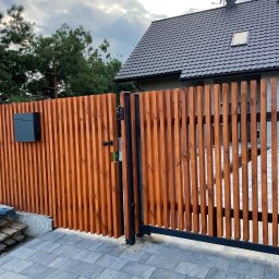 Dawid Karolewski - Nowoczesna brama wjazdowa z drewnianymi lamelami w ciepłym odcieniu brązu, czarna skrzynka na listy, szara kostka brukowa i dom w tle pod pochmurnym niebem.